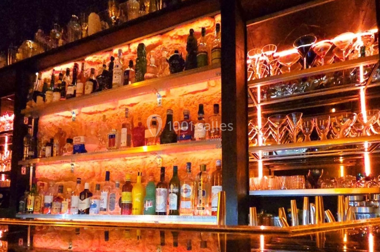 Étagères illuminées d’un bar avec bouteilles d’alcool variées et verres disposés sur un comptoir en bois.