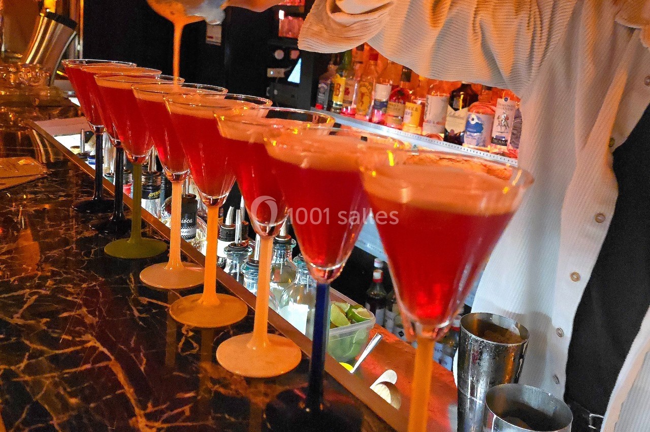 Une rangée de cocktails rouges alignés sur un bar, avec une personne versant un liquide dans l'un des verres.