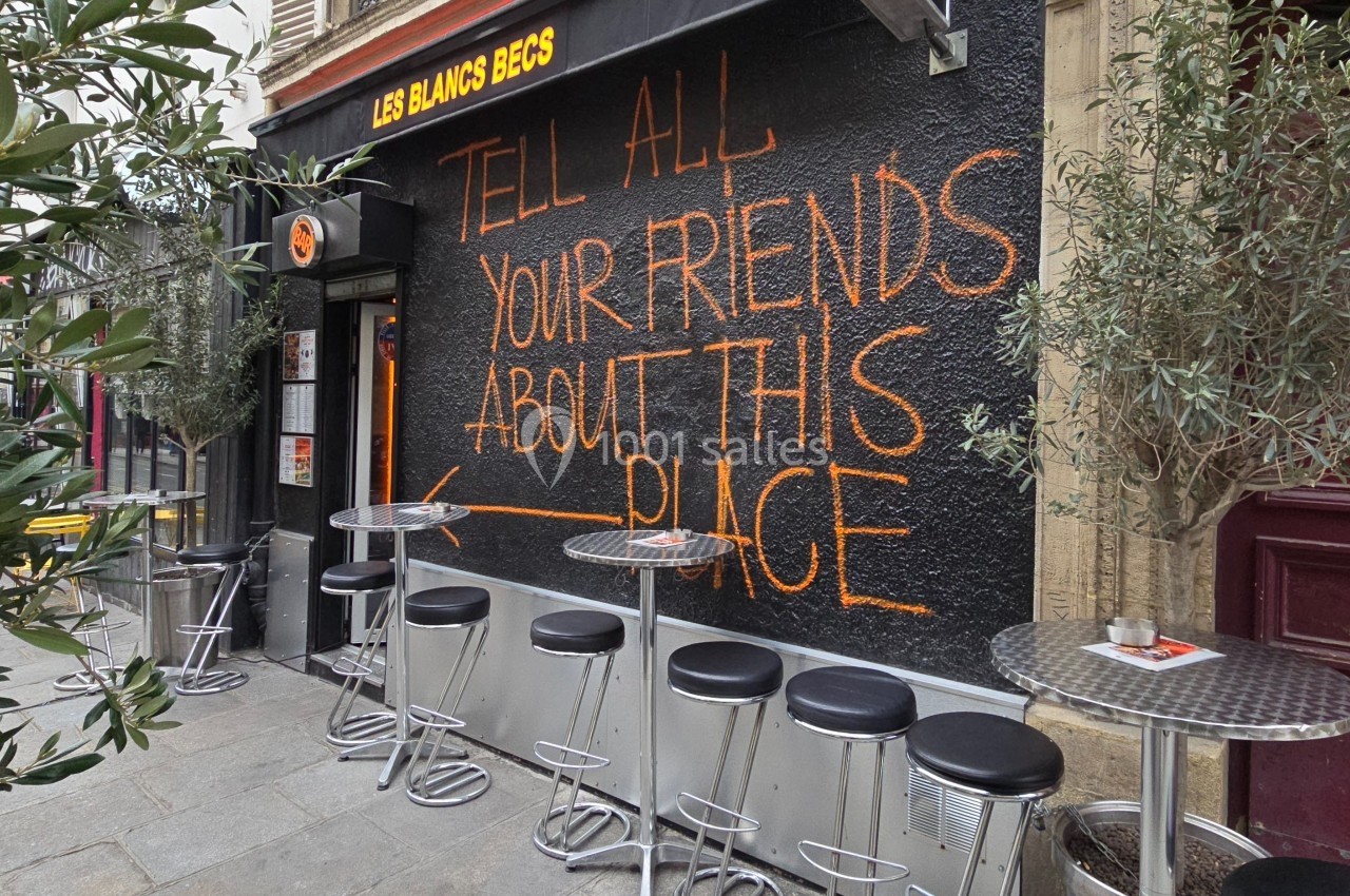 Façade d'un café avec des tables hautes et un mur noir portant une inscription orange manuscrite.