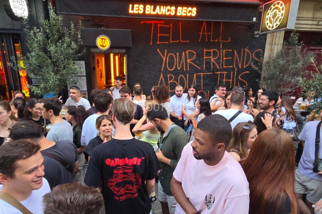 Foule rassemblée devant un bar animé, avec une enseigne lumineuse et un message peint sur le mur.