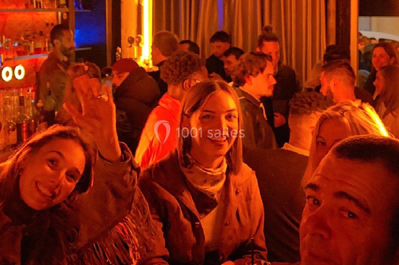 Groupe de personnes souriantes dans un bar animé, éclairé par des lumières chaudes et entouré d'autres clients.
