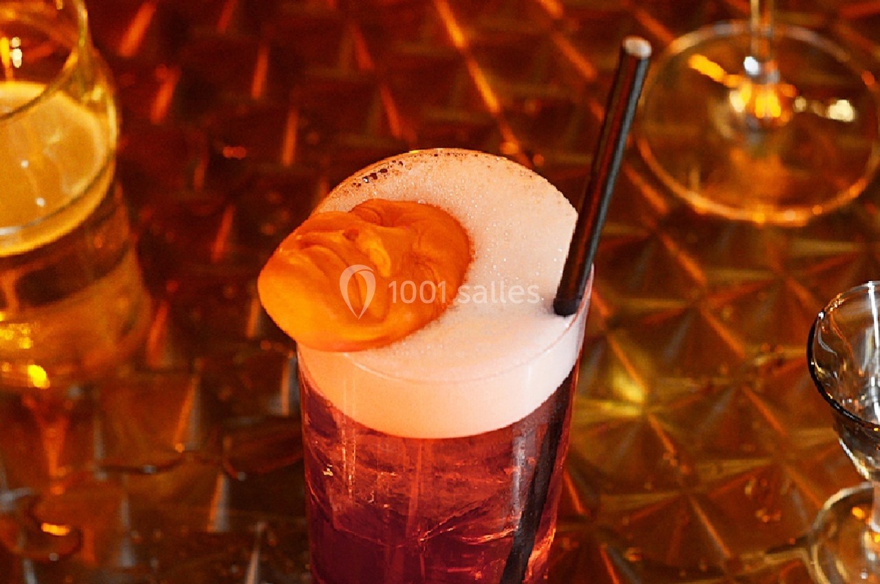 Verre de cocktail rouge avec mousse blanche, décoré d'une figure sculptée orange, posé sur une table texturée.