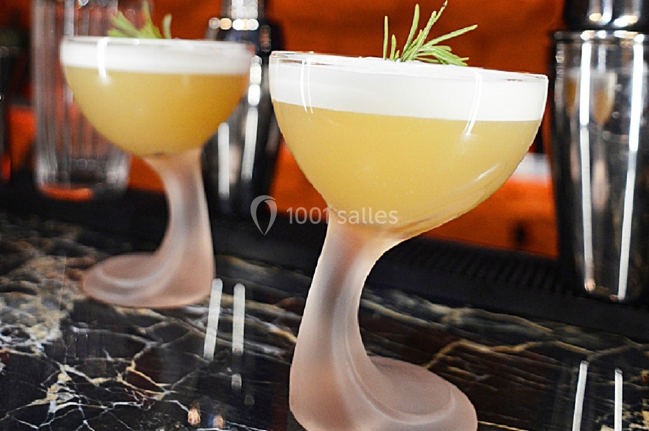Deux cocktails jaunes avec mousse blanche, garnis de romarin, servis dans des verres au design incurvé sur un bar en marbre.