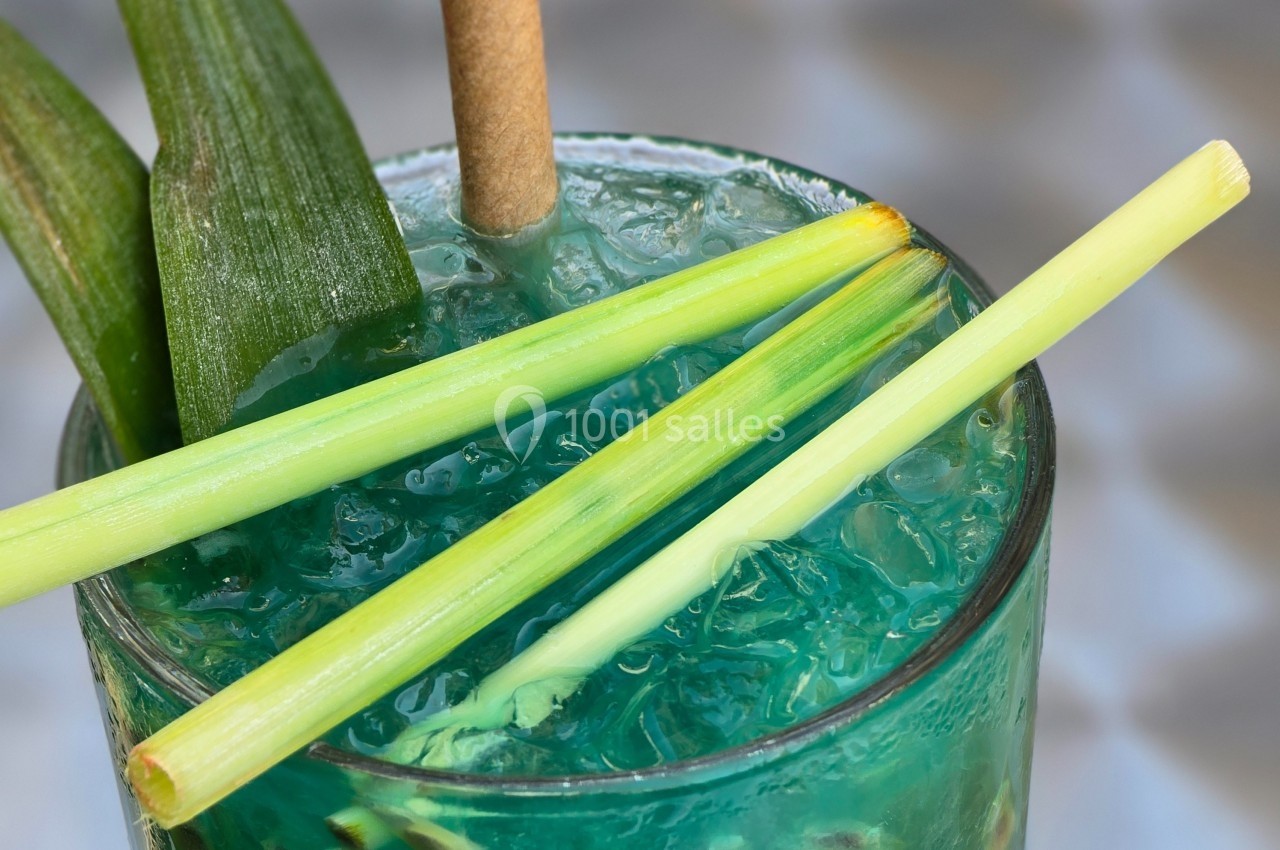 Verre de cocktail bleu avec glaçons, décoré de feuilles et de tiges de citronnelle.