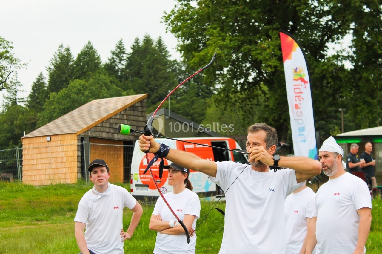 Un homme tire à l'arc en plein air, entouré de personnes en tenue blanche, avec des bâtiments et des arbres en arrière-plan.