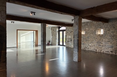 Salle spacieuse avec murs en pierre, poutres en bois apparentes et sol en carrelage gris, éclairée par des fenêtres.