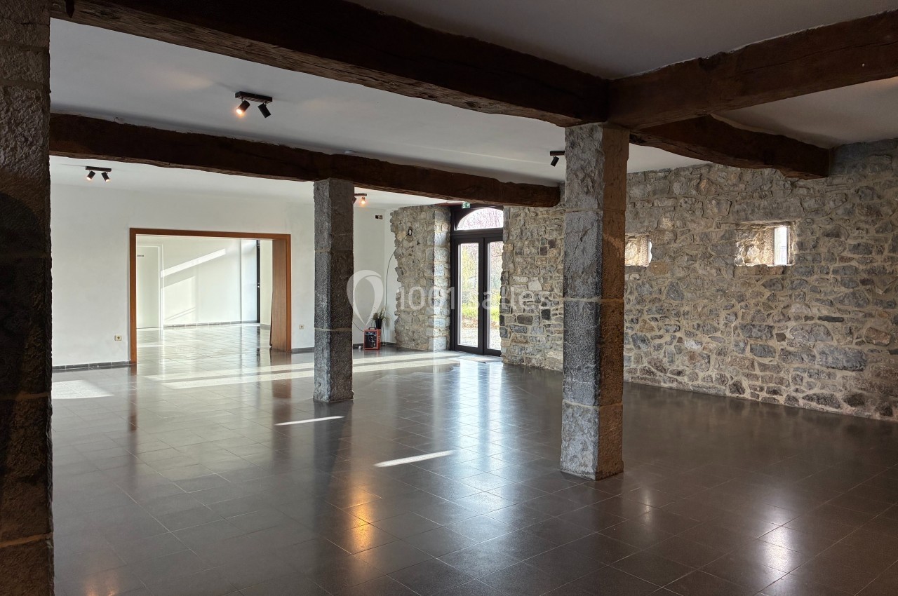 Salle spacieuse avec murs en pierre, poutres en bois apparentes et sol en carrelage gris, éclairée par des fenêtres.