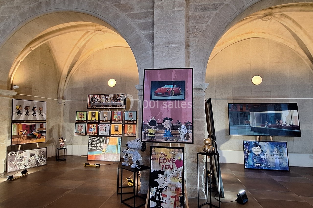 Exposition d'art contemporain dans une salle voûtée en pierre, avec peintures et sculptures disposées sur des supports.