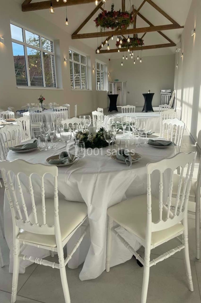 Salle de réception lumineuse avec tables rondes dressées, chaises blanches et poutres apparentes au plafond.