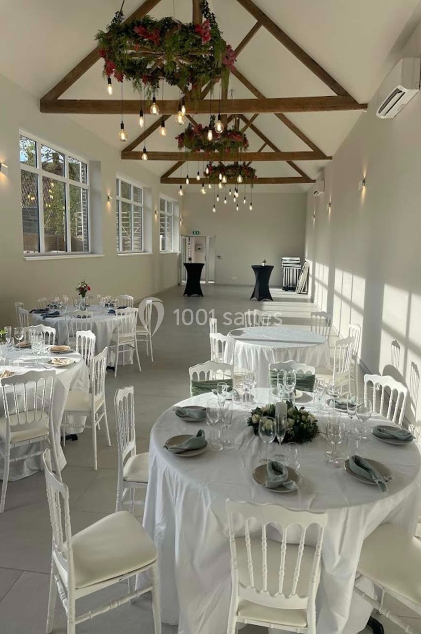Salle lumineuse avec tables rondes dressées, chaises blanches, poutres apparentes et décorations suspendues.