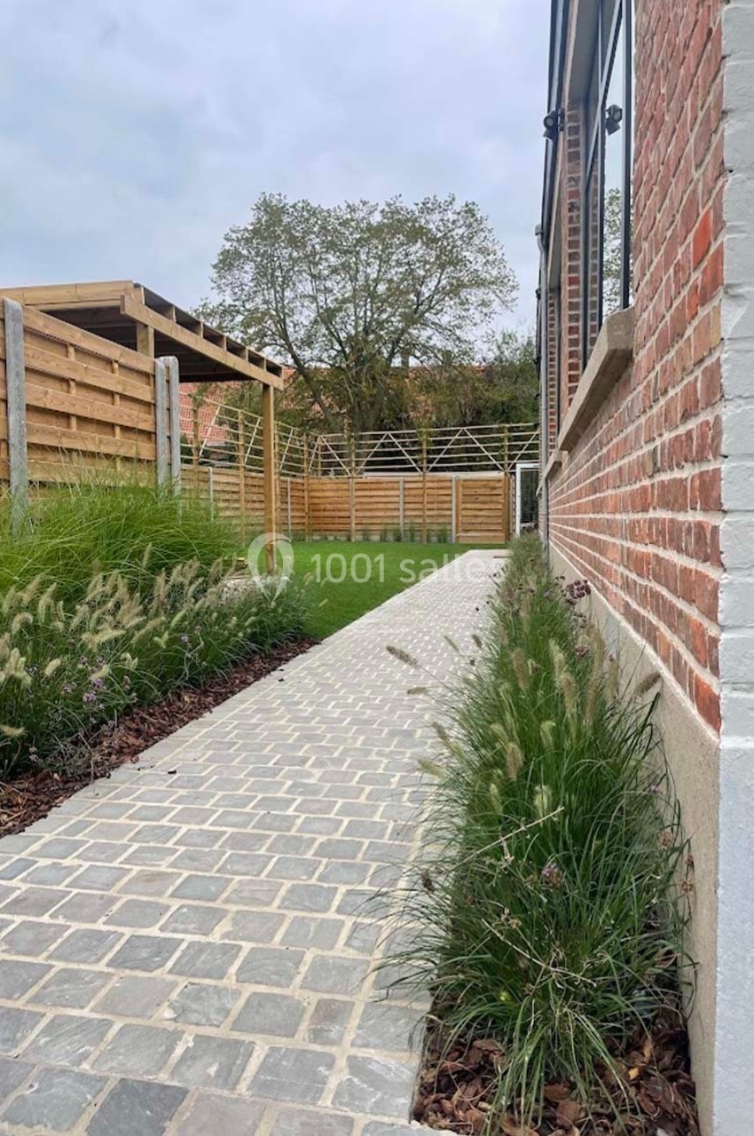 Allée pavée bordée de plantes, menant à un jardin avec pelouse et pergola en bois, près d'un mur en briques.