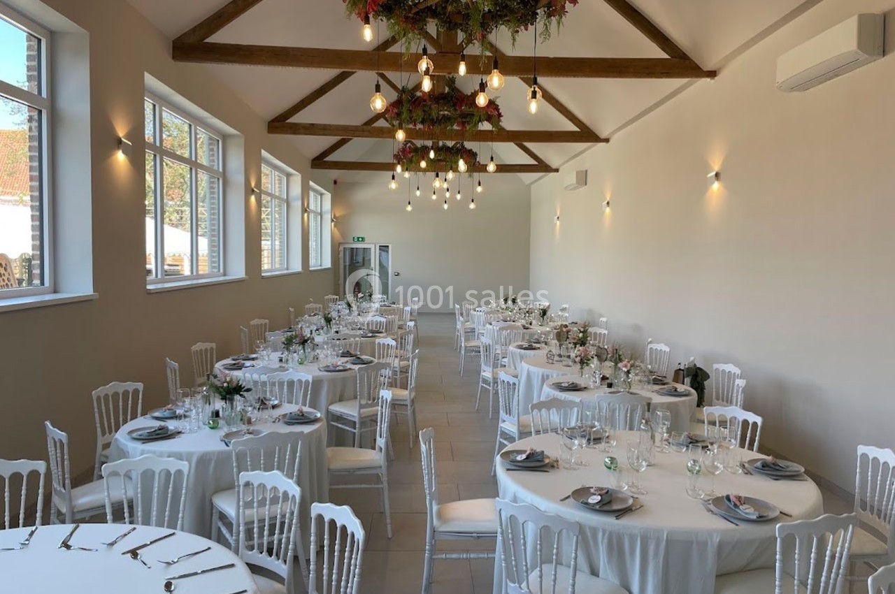 Salle de réception lumineuse avec tables rondes dressées, chaises blanches et décorations florales suspendues.