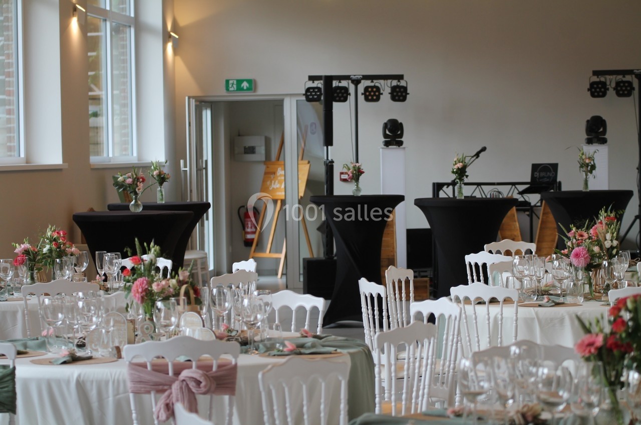 Salle de réception décorée pour un événement, avec tables dressées, chaises blanches et arrangements floraux.