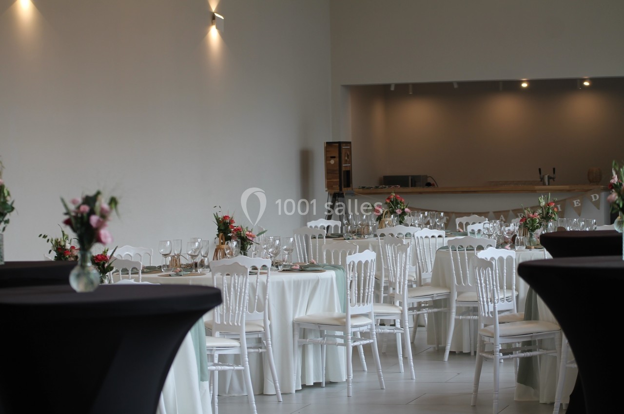 Salle de réception décorée avec des tables rondes nappées de blanc, chaises blanches et arrangements floraux.
