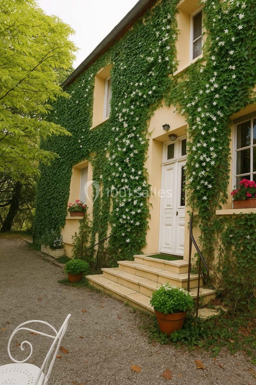 Façade d'une maison recouverte de lierre avec des marches en pierre, des pots de fleurs et un arbre à gauche.