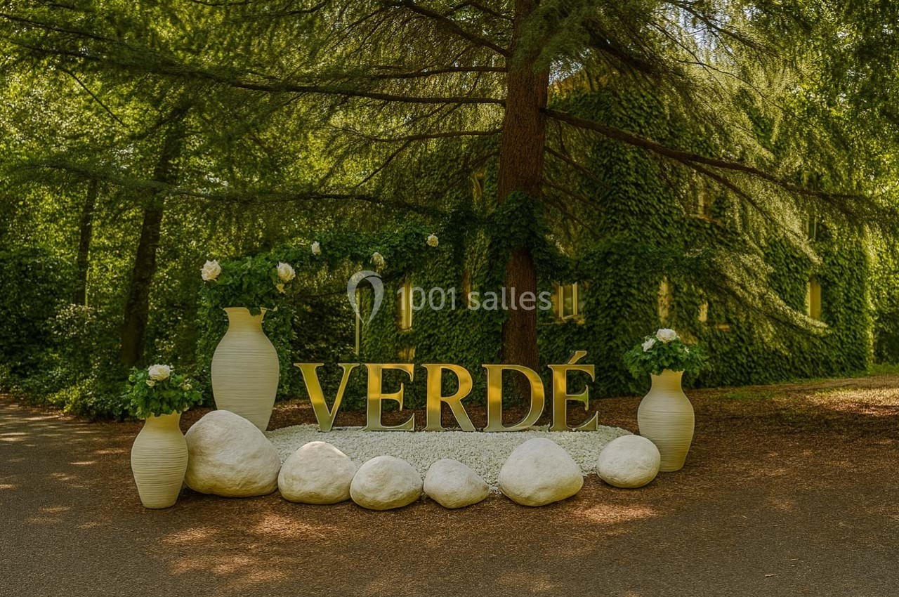 Installation décorative avec des vases blancs, des plantes et des lettres formant ’VERDÉ’, entourée de pierres blanches…