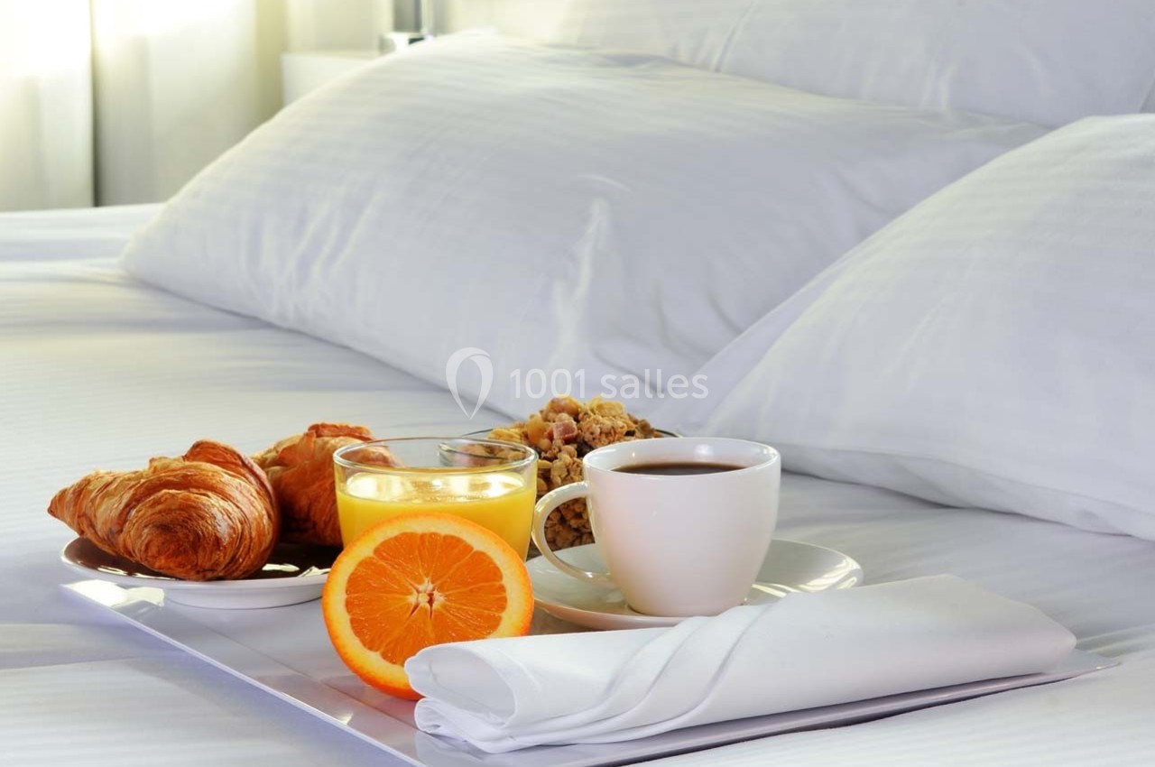 Petit-déjeuner avec croissants, jus d'orange, café et céréales servi sur un plateau posé sur un lit blanc.