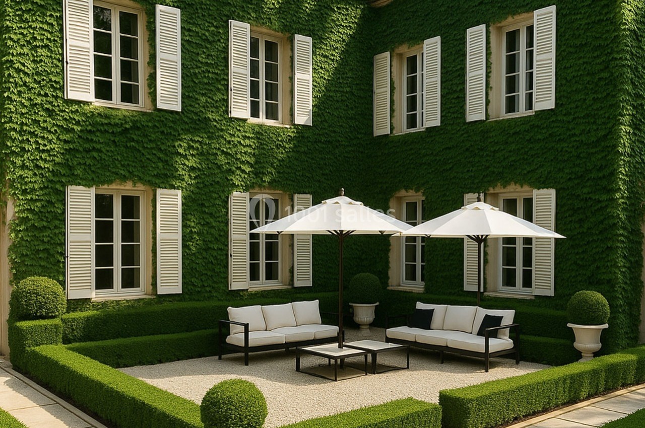 Façade d'une maison recouverte de lierre avec une cour aménagée, des parasols blancs et des haies taillées.