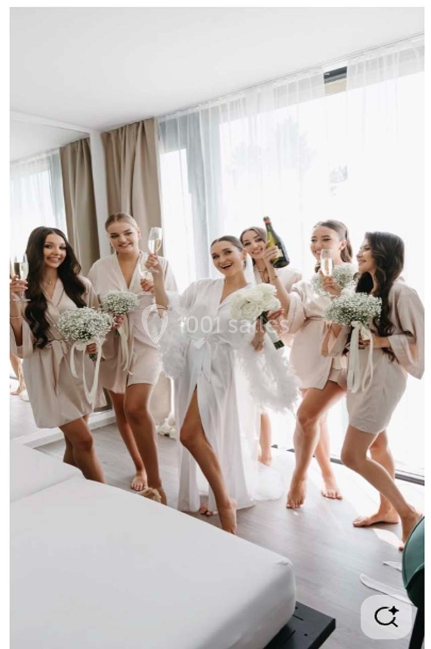 Un groupe de femmes en peignoirs, tenant des coupes de champagne et des bouquets, dans une chambre lumineuse.
