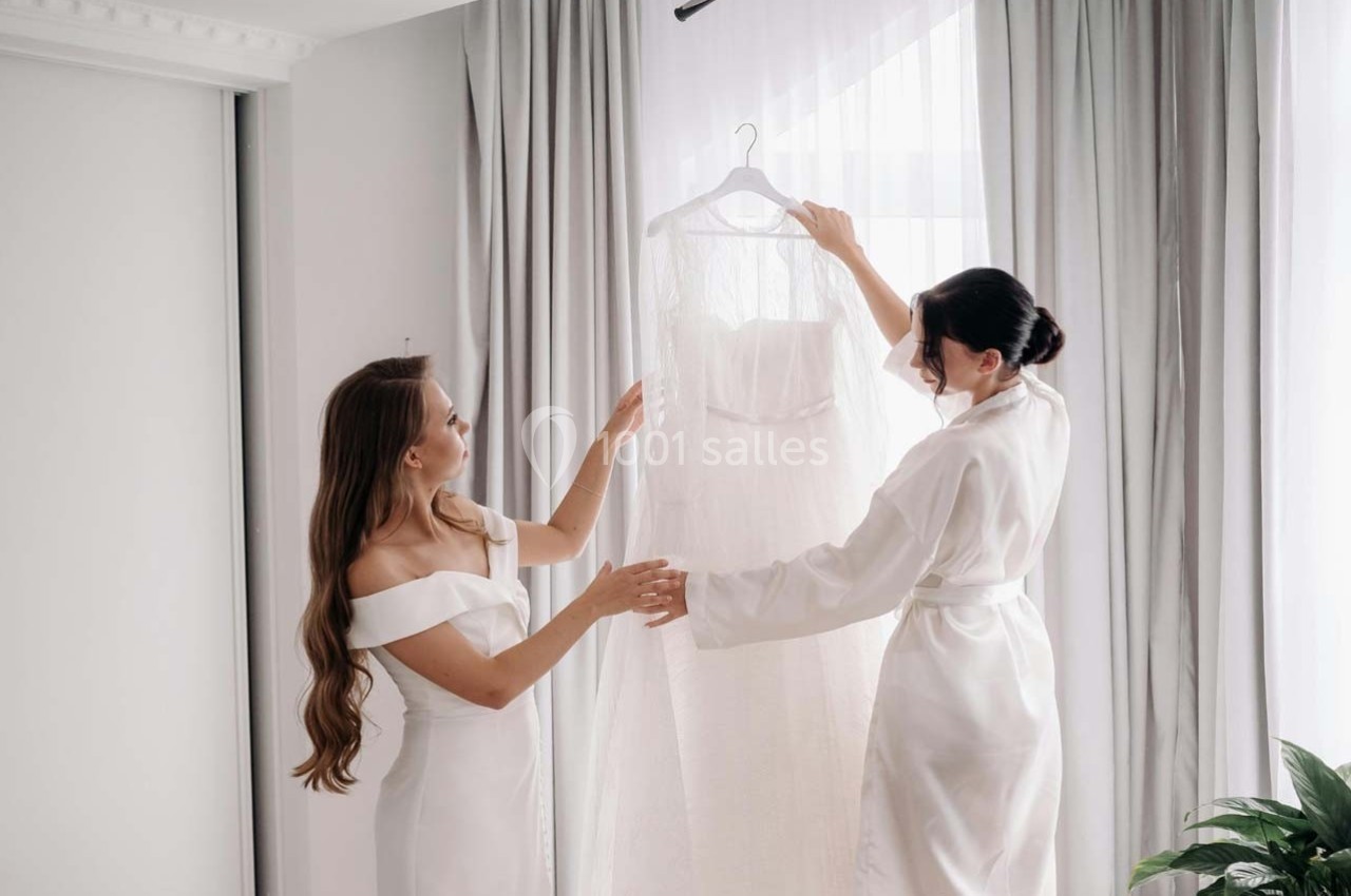 Deux femmes en robes blanches tiennent une robe de mariée près d'une fenêtre avec des rideaux clairs.