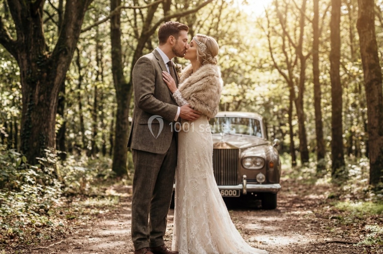 Un couple en tenue de mariage s'embrasse dans une forêt, avec une voiture ancienne en arrière-plan.