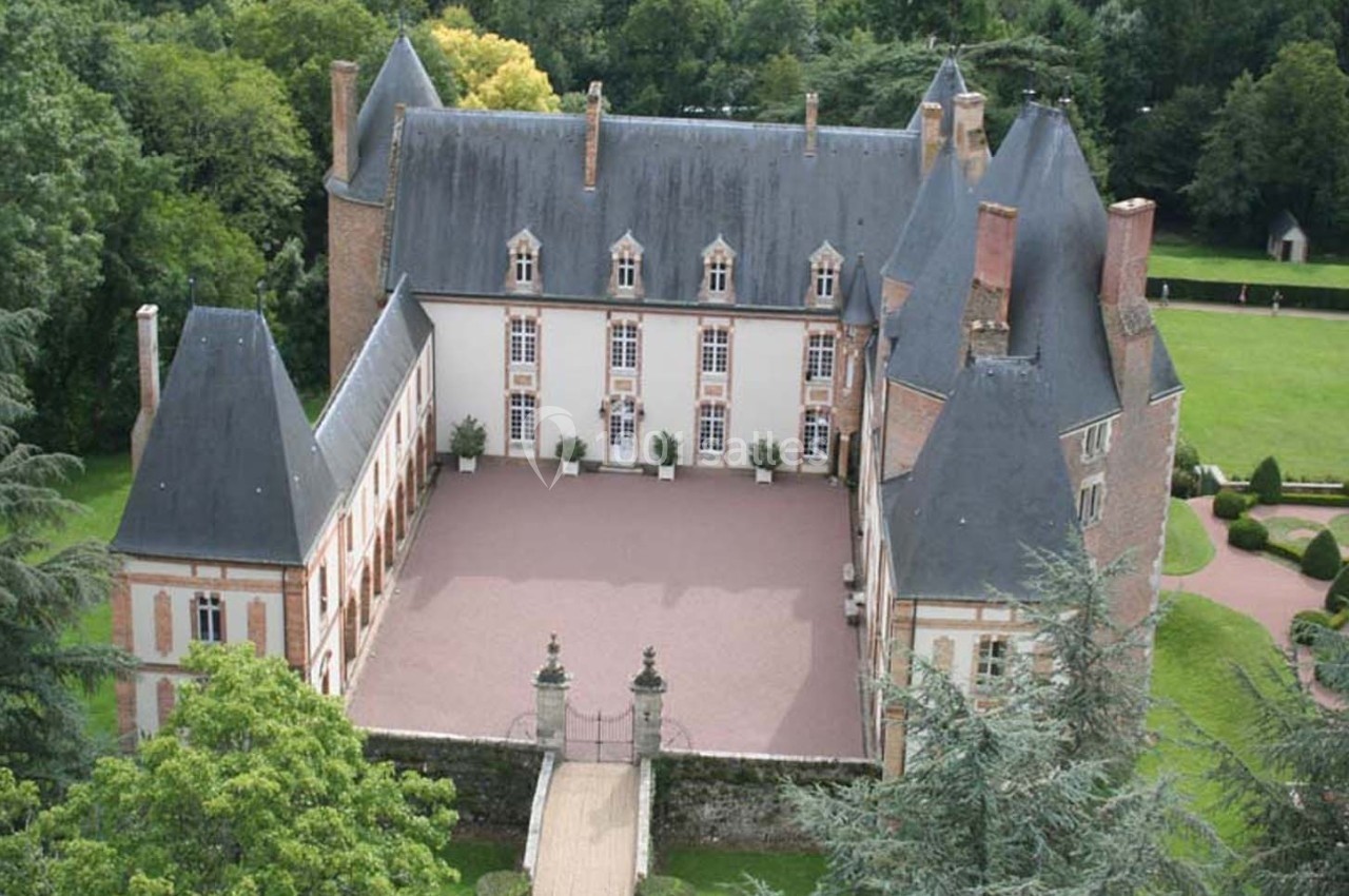 Vue aérienne d'un château entouré de jardins et d'arbres, avec une cour centrale pavée.