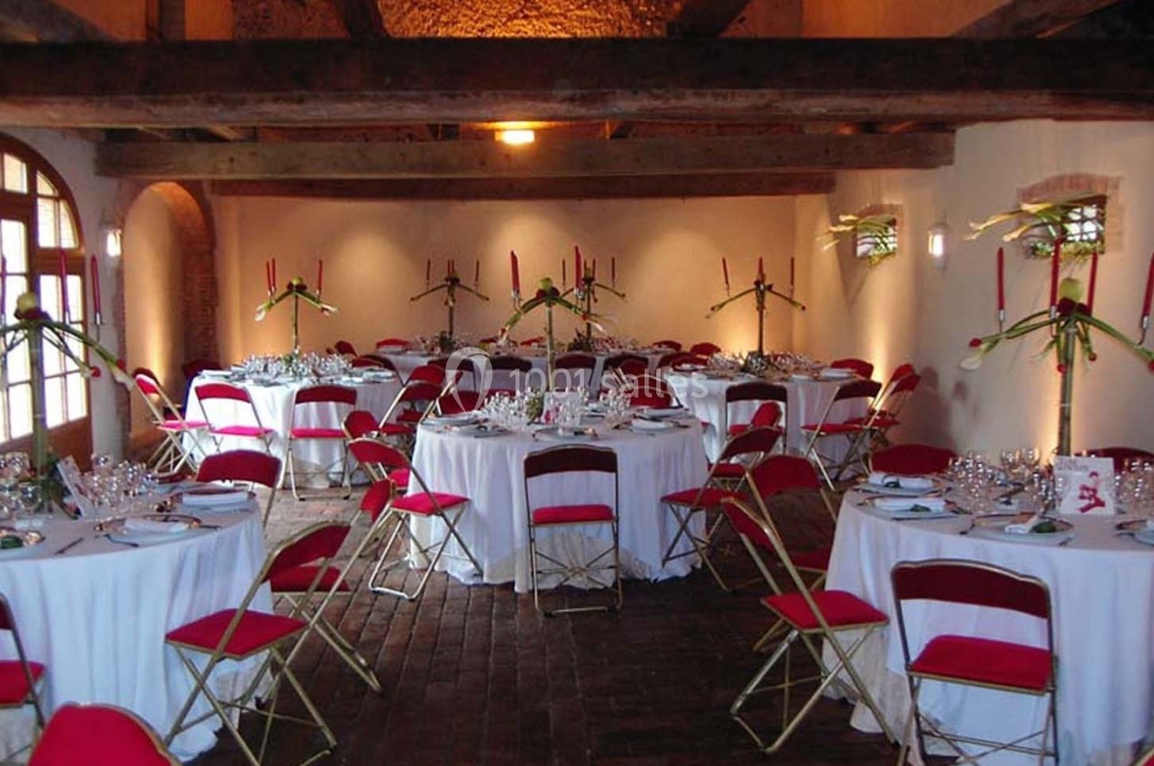 Salle de réception avec tables rondes dressées, nappes blanches, chaises rouges et décorations florales.
