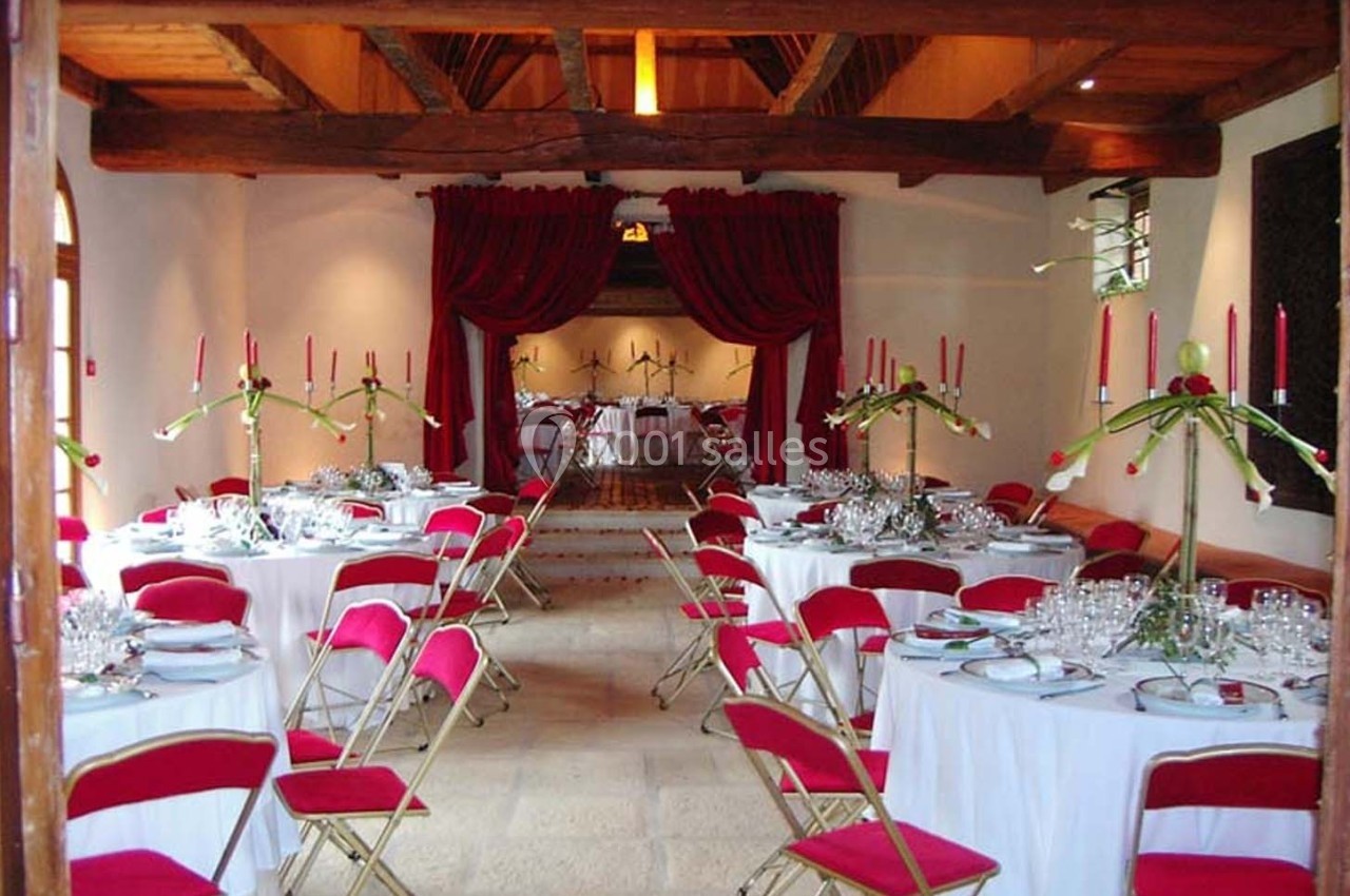 Salle de réception décorée avec des tables rondes, nappes blanches, chaises rouges et chandeliers ornés de bougies.