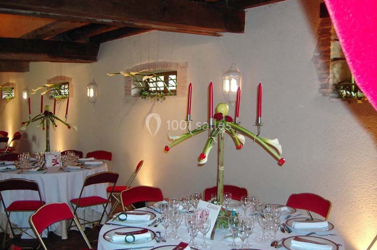 Salle de réception décorée avec des tables rondes, nappes blanches, chaises rouges et centre de table floral avec bougies.