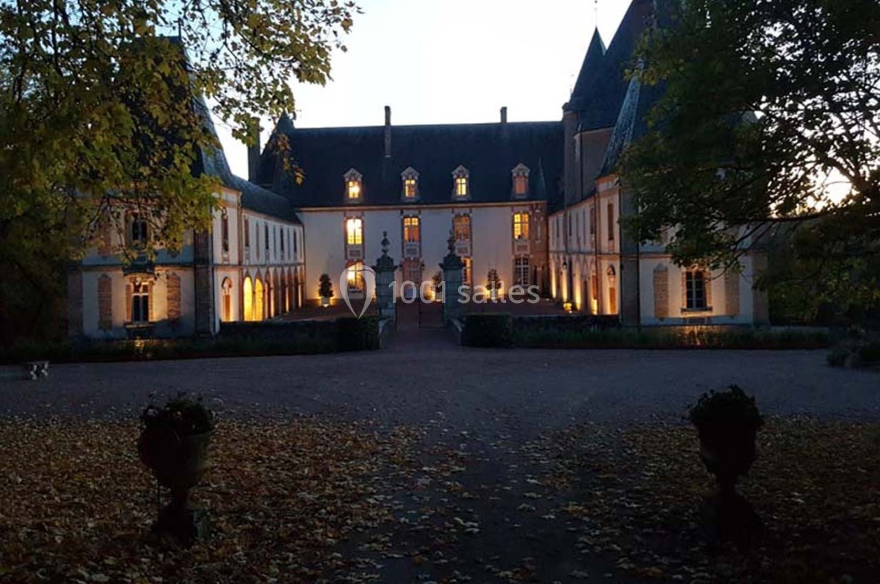 Façade d'un château éclairé à la tombée de la nuit, entouré d'arbres et d'une cour jonchée de feuilles.