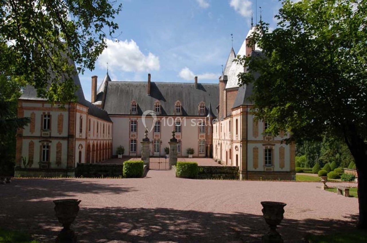 Façade d'un château entouré d'arbres, avec une cour pavée et un ciel partiellement nuageux.