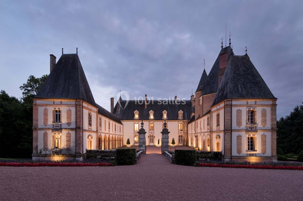 Façade d'un château éclairé au crépuscule, entouré de jardins et d'une allée gravillonnée.