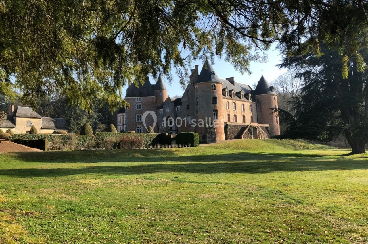 Château en briques rouges entouré de verdure, avec des tours et un grand parc arboré sous un ciel dégagé.