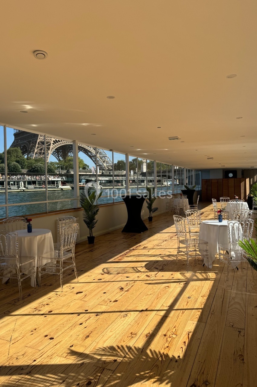 Salle lumineuse avec parquet, tables dressées, grandes baies vitrées offrant une vue sur la Tour Eiffel et la Seine.