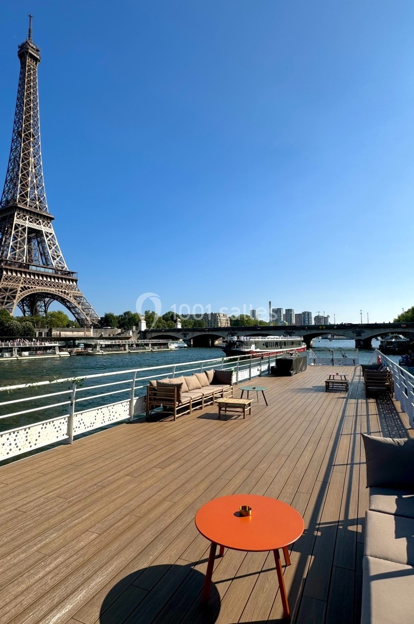 Vue d'une terrasse en bois sur une péniche avec mobilier, face à la tour Eiffel et la Seine par temps ensoleillé.