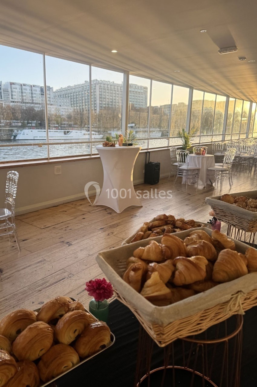 Salle lumineuse avec vue sur la Seine, tables hautes décorées et paniers de viennoiseries au premier plan.