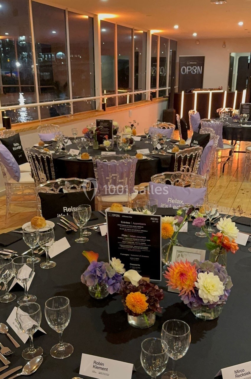 Tables élégamment dressées avec nappes noires, fleurs colorées et menus, dans une salle avec vue sur l'eau en soirée.