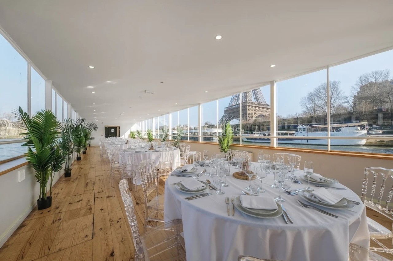 Salle de réception lumineuse avec vue sur la Tour Eiffel, tables dressées et grandes baies vitrées donnant sur la Seine.
