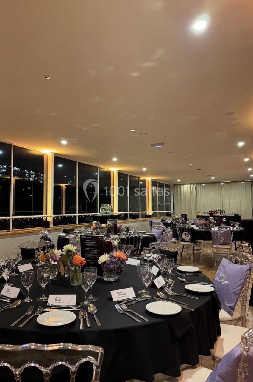 Salle élégamment décorée pour un dîner, avec tables dressées, nappes noires, chaises transparentes et vue nocturne sur la…