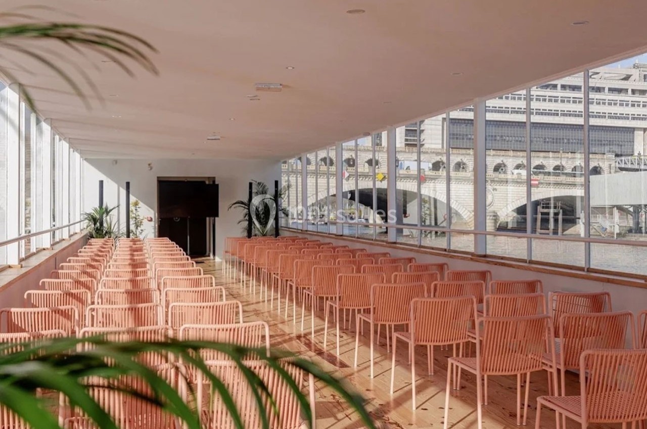 Salle lumineuse avec des chaises roses alignées, grandes baies vitrées donnant sur un bâtiment et un pont au bord de l'eau.