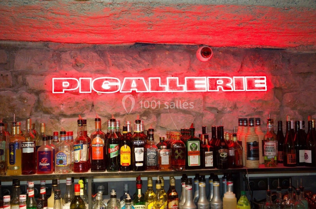 Bar avec un mur en pierre, des bouteilles d'alcool alignées et une enseigne lumineuse rouge indiquant ’Pigallerie’.