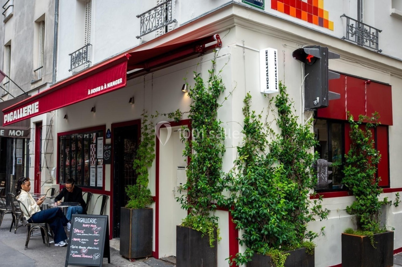 Façade d'un café avec terrasse, plantes grimpantes et enseigne rouge, situé à un coin de rue en ville.