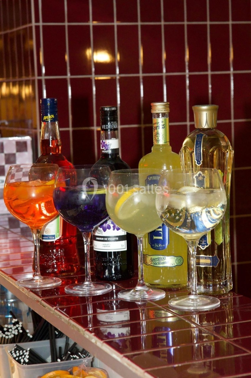 Verres de cocktails colorés alignés devant des bouteilles d'alcool sur un comptoir carrelé rouge.