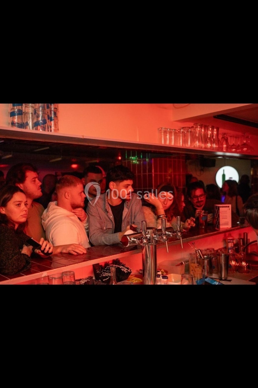 Des personnes discutent et attendent au comptoir d’un bar éclairé par une lumière rouge tamisée.