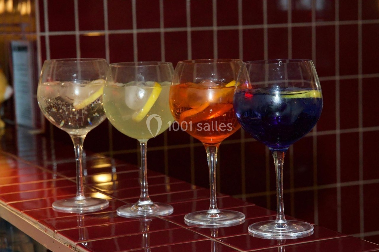 Quatre verres à pied contenant des cocktails colorés avec des tranches de citron, alignés sur un comptoir carrelé rouge.