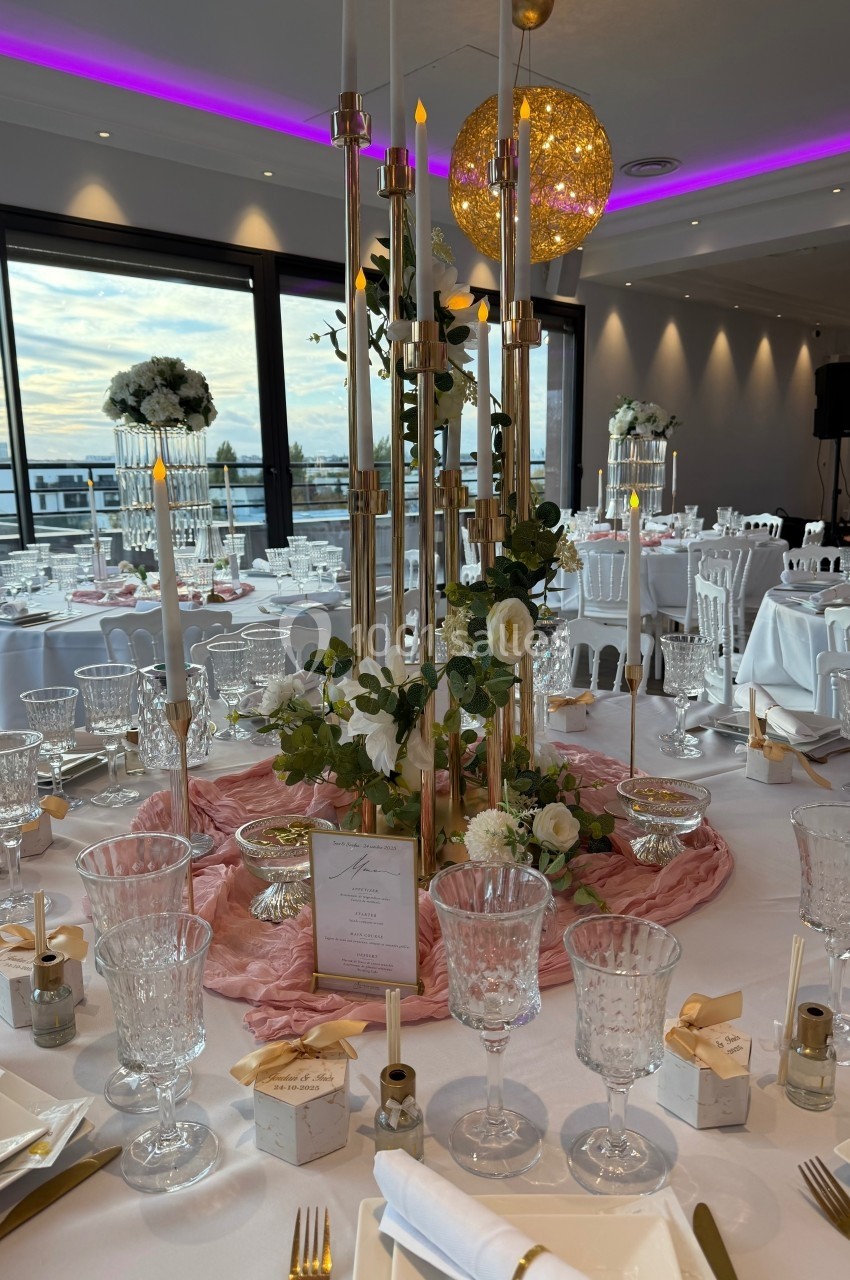 Salle de réception élégamment décorée avec des tables dressées, des chandeliers dorés et des fleurs blanches.