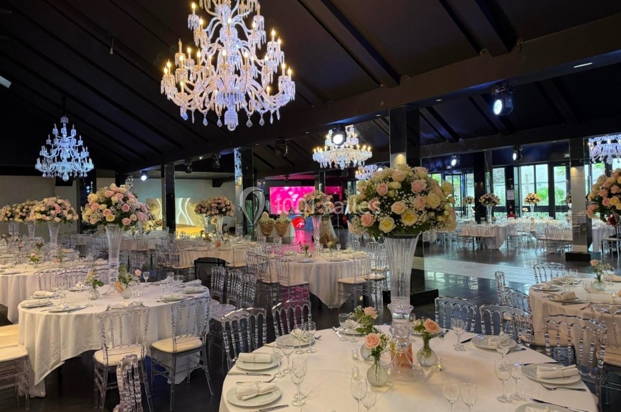 Salle de réception élégante avec tables dressées, chaises transparentes, chandeliers et décorations florales.