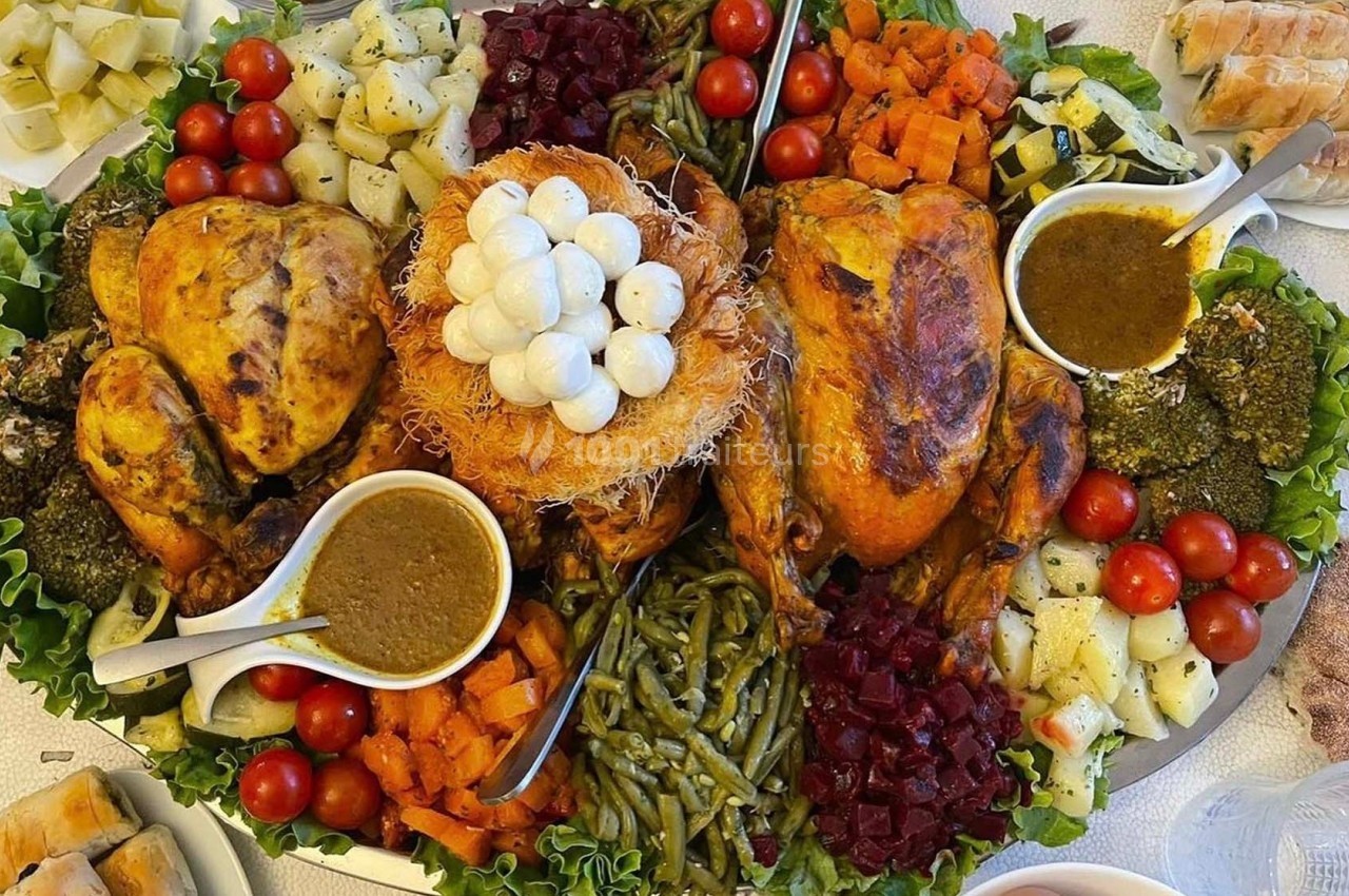Plateau de repas festif avec poulets rôtis, légumes variés, sauces et garnitures disposés sur des feuilles de salade.