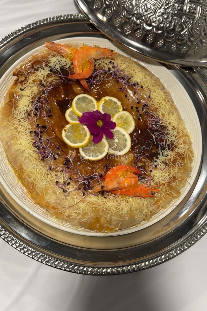 Plat de présentation avec une pastilla garnie de crevettes, citron, fleurs comestibles et décorée de vermicelles dorés.
