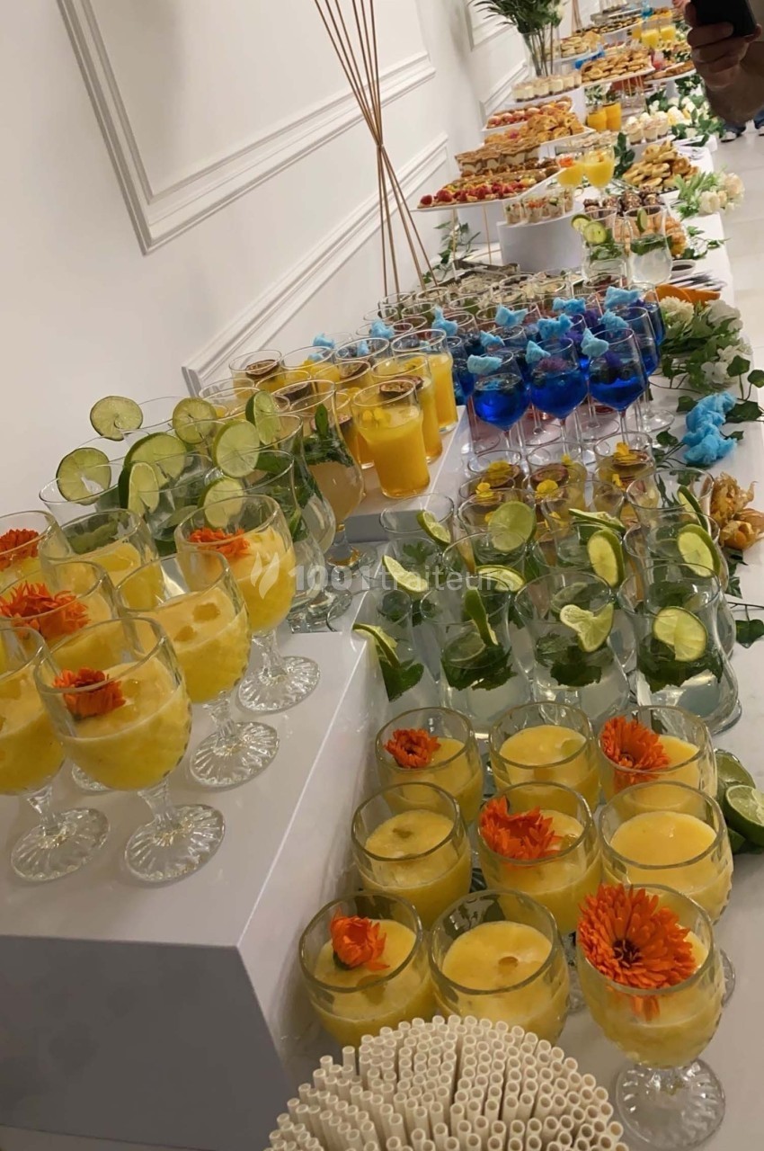Buffet de boissons colorées avec cocktails garnis de fruits, fleurs et glaçons, disposés sur une table blanche.